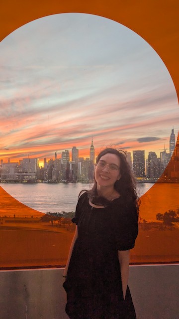 Shelly Xiong in front of a circular window framing the NYC skyline at sunset